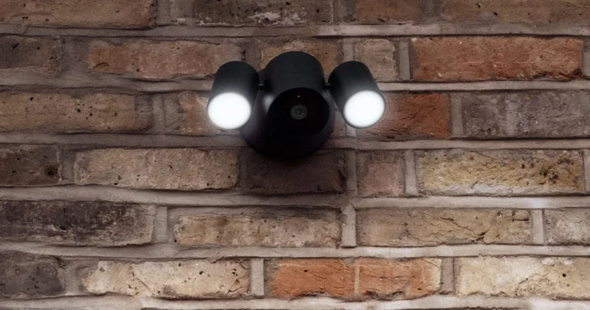 A black, dual-head outdoor security floodlight with a central camera lens mounted on a rustic brick wall.