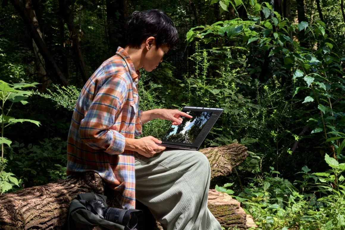 Apple’ın İlk Dokunmatik OLED MacBook Pro’su Tanıtıldı 20 A person wearing a plaid shirt sits on a fallen log in a forest, using a touchscreen laptop while a camera rests beside them.