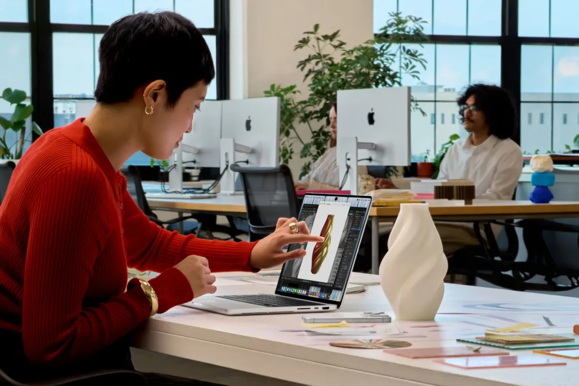 Apple’ın İlk Dokunmatik OLED MacBook Pro’su Tanıtıldı 21 A person in a red sweater sits at a desk, using their finger to interact with a laptop screen displaying a 3D design, while others work in the background.