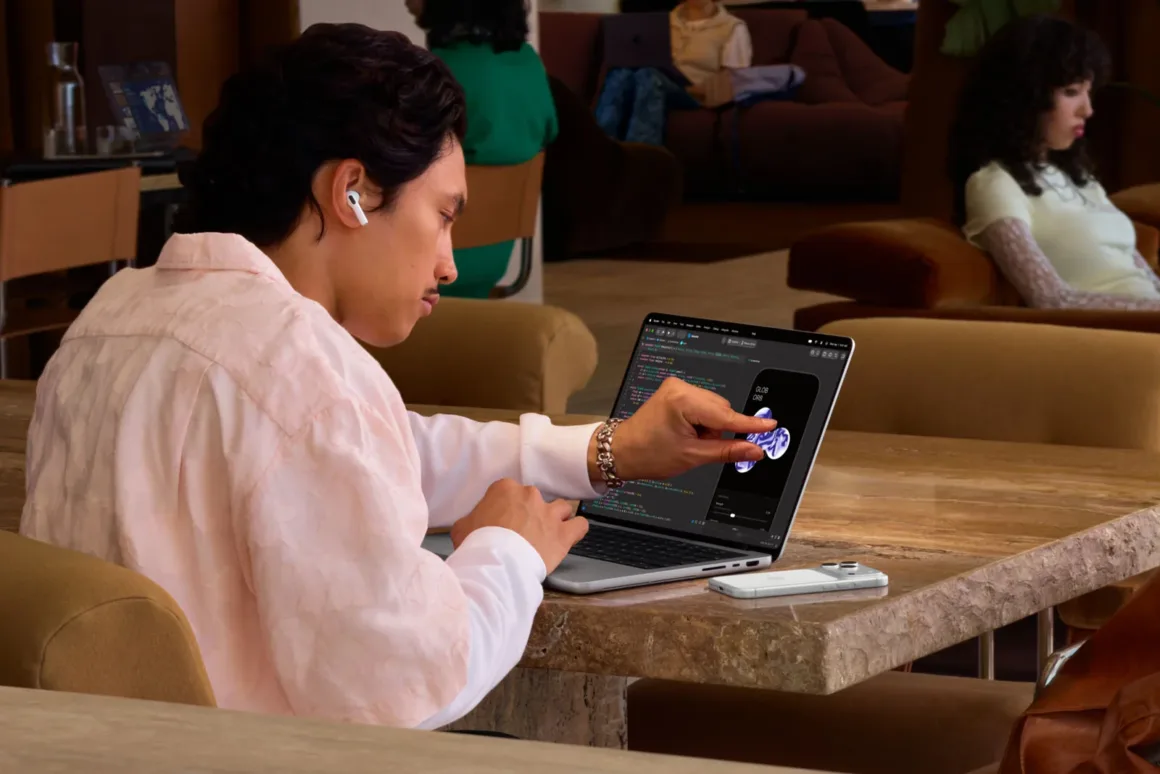 Apple’ın İlk Dokunmatik OLED MacBook Pro’su Tanıtıldı 22 A person with dark hair and a light pink shirt sits at a wooden table, touching the screen of an open laptop while wearing an earbud.