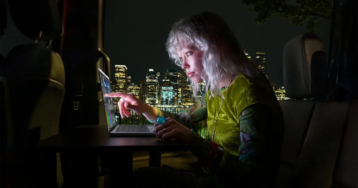 A person with blonde hair sits in the back of a vehicle at night, using a laptop while looking at a city skyline.
