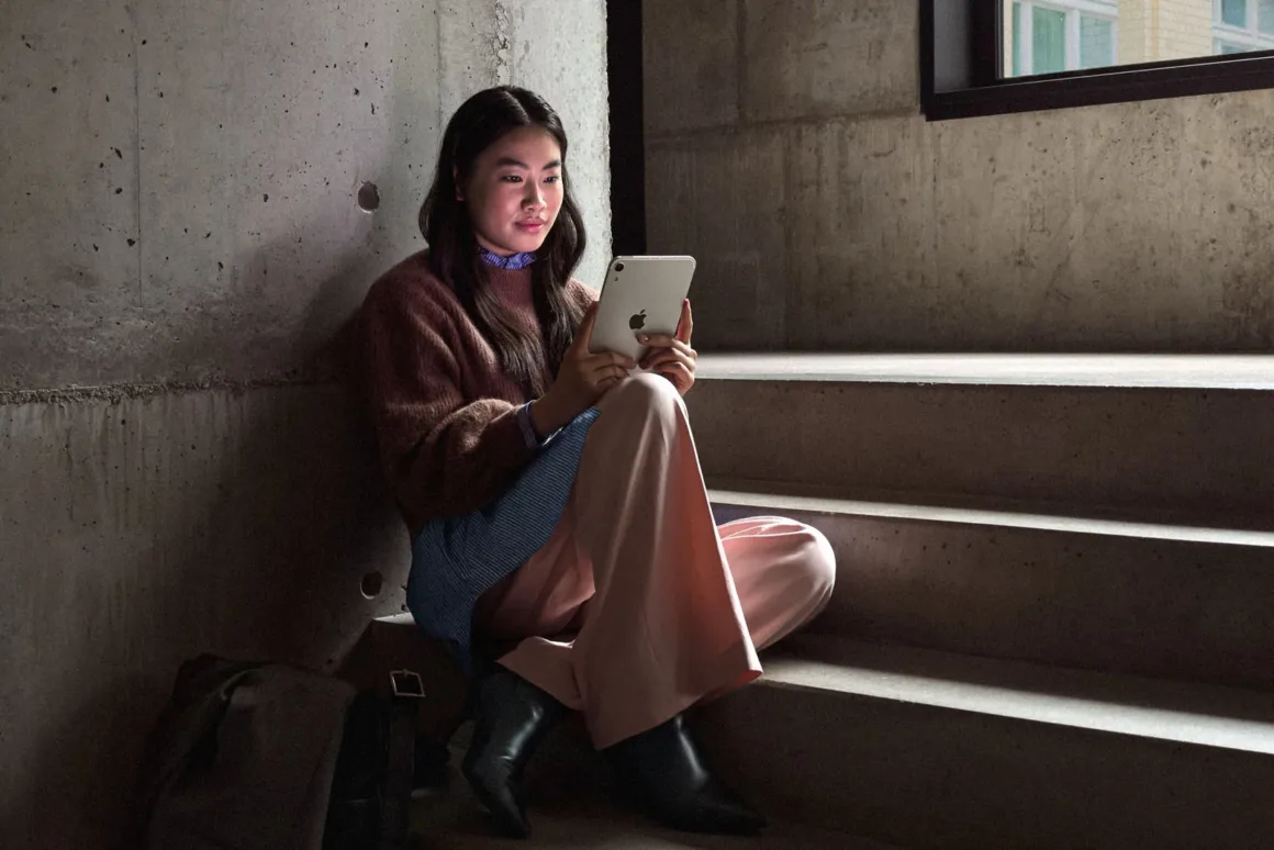 Yeni iPad mini Tanıtıldı: OLED Ekran, A20 Pro ve Daha İnce Tasarım 23 Uzun koyu saçlı genç bir kadın kahverengi kazak ve açık renk pantolonla beton basamaklarda otururken elindeki tableti kullanıyor.