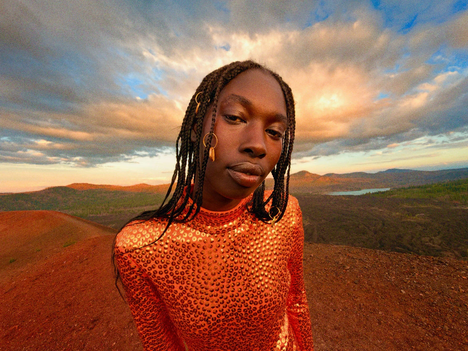 Apple iPhone Duo: İlk Katlanabilir iPhone 7,6 İnç Ekranla Geldi 29 A person with braided hair and gold hair accessories wears a shimmering orange sequined top while standing on a rocky, reddish landscape at sunset.