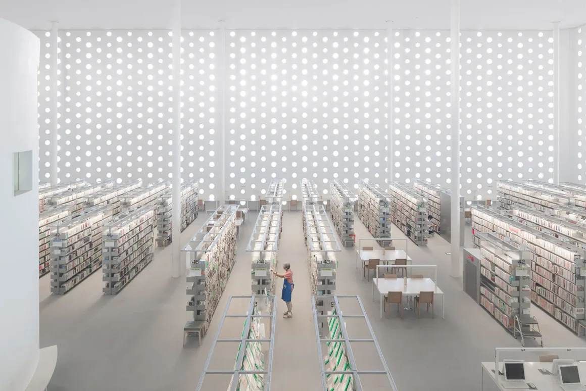 Architecture Photography MasterPrize 2026 Kazananları Açıklandı 20 Yüksek açıdan görülen aydınlık ve minimalist kütüphanede kitap rafları sıralanıyor; bir kişi koridorda dururken duvarda tekrarlayan dairesel açıklıklar görülüyor.