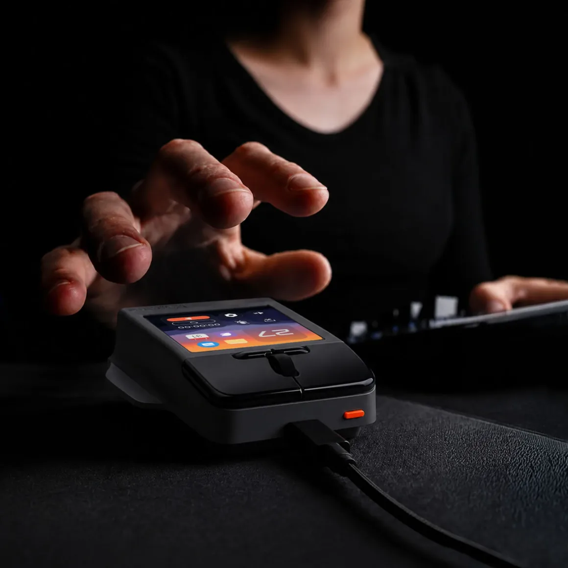 Super Mouse Tanıtıldı: Dokunmatik Ekranlı Fare Üretkenlik Merkezi Olmayı Hedefliyor 27 A person's hand reaches toward a small, grey, rectangular device with a touchscreen display and a wired connection, resting on a dark surface.