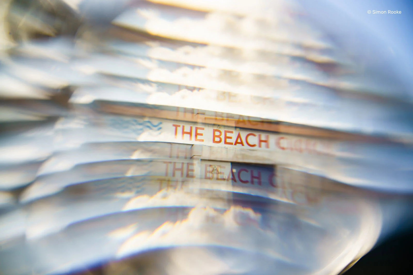 Lensbaby Omni Color II ve Omni Crystal II ile Çekim Sırasında Yaratıcı Efektler 27 A distorted, multi-layered view of a sign reading "THE BEACH CLUB" seen through a glass lens or prism.