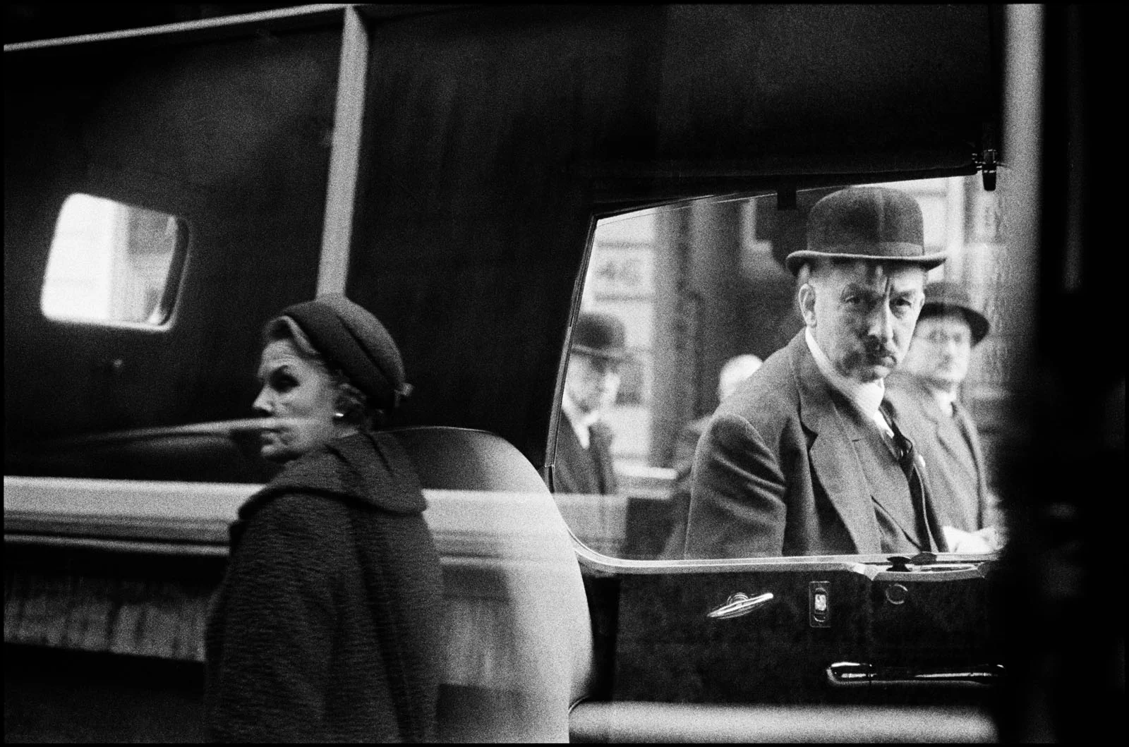 Magnum Fact/Fiction Square Print Sale 26 Ekim’de Başlıyor 30 A woman in a hat stands in the foreground while a man in a bowler hat looks out from the window of a vehicle.