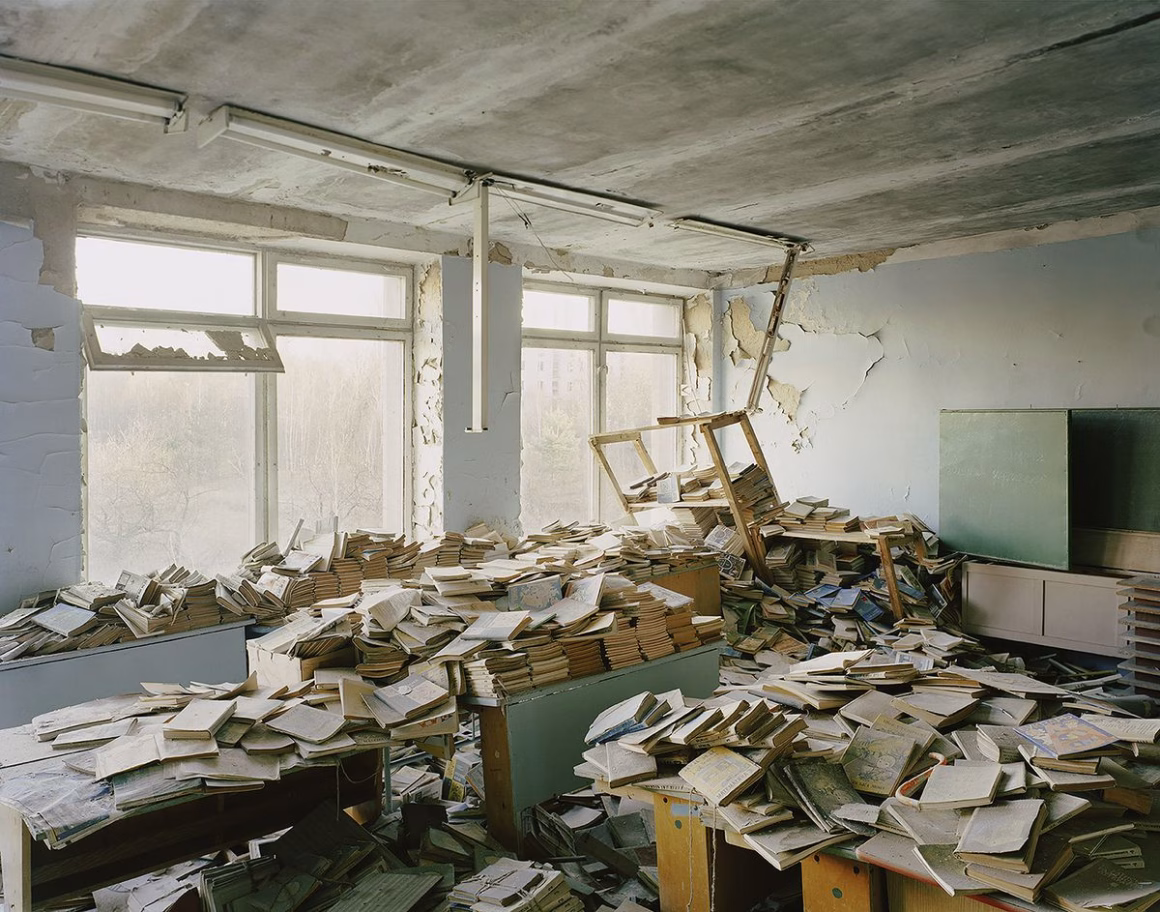 Pripyat'taki terk edilmiş bir okul kütüphanesi; tozlu kitap yığınları masaların ve zeminin üzerine dağılmış, duvarların boyaları dökülmüş ve tavandaki lamba sarkıyor.