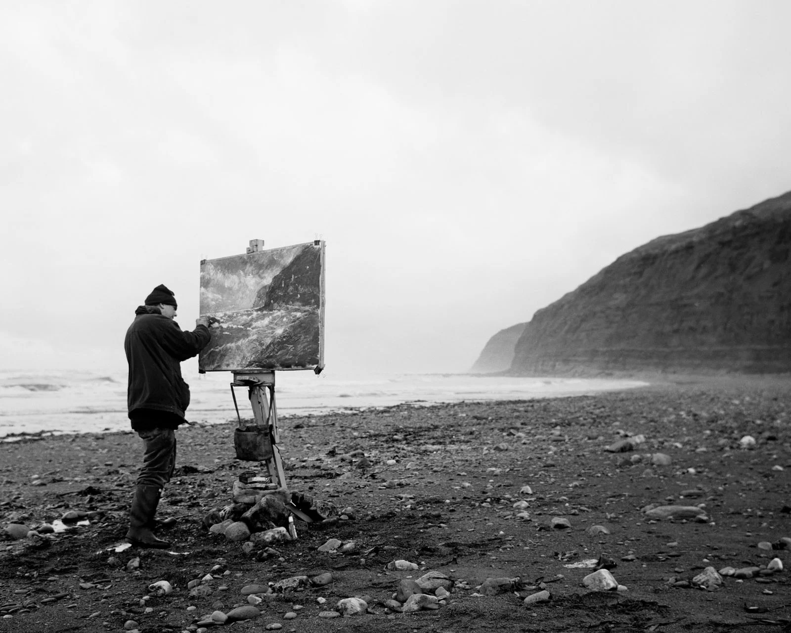 Magnum Fact/Fiction Square Print Sale 26 Ekim’de Başlıyor 29 A person wearing a beanie and dark jacket paints a coastal landscape on an easel set up on a rocky beach.