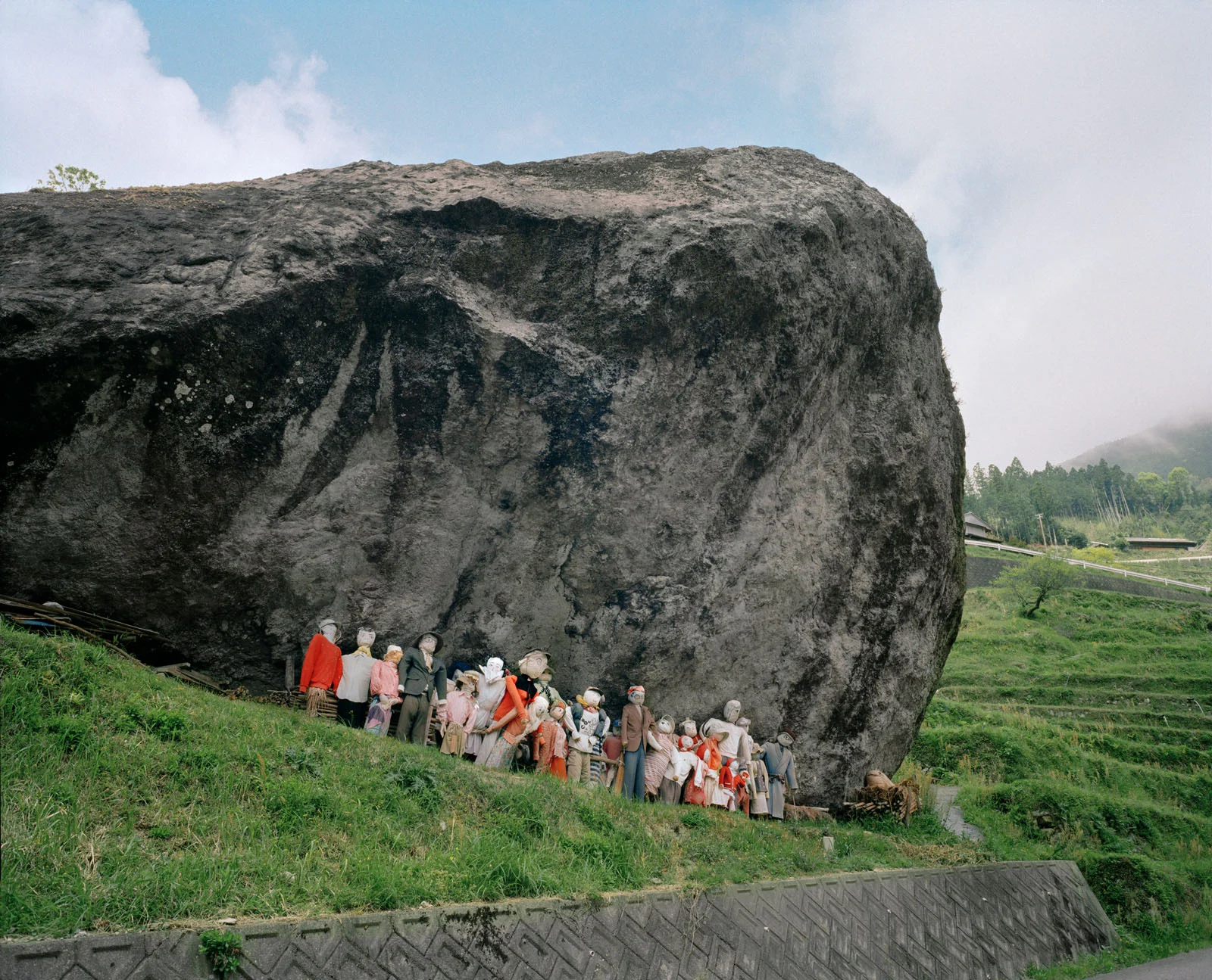Magnum Fact/Fiction Square Print Sale 26 Ekim’de Başlıyor 26 A large, dark boulder sits on a grassy hillside, with a row of human-sized scarecrows dressed in various clothes standing at its base.