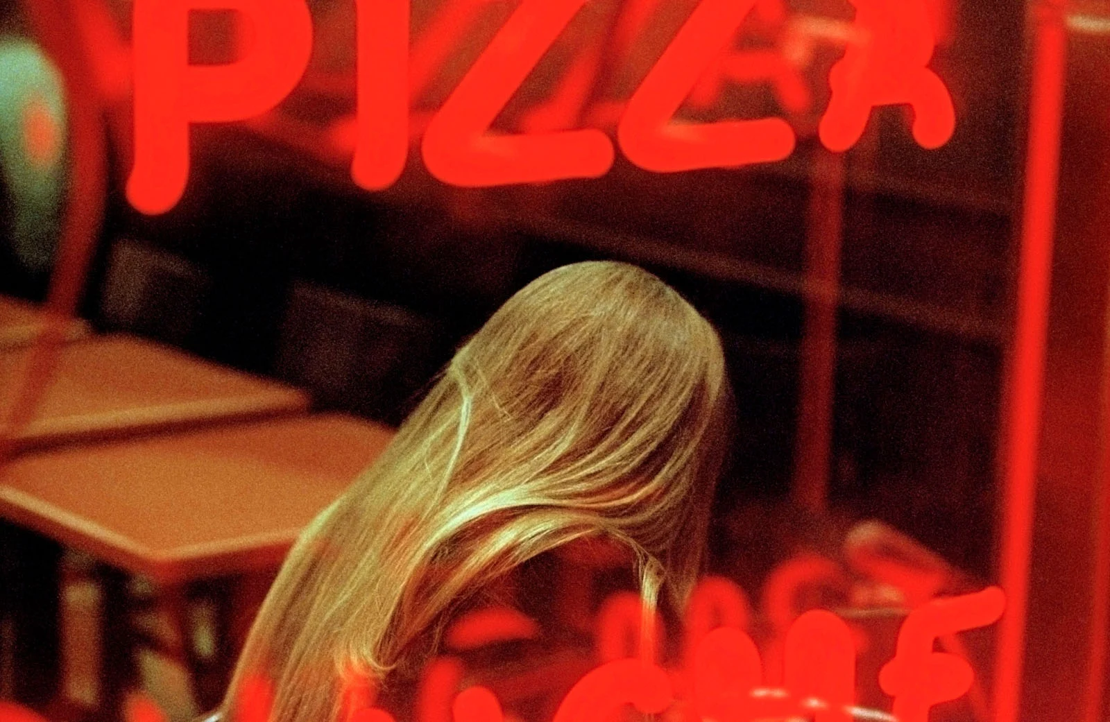 Magnum Fact/Fiction Square Print Sale 26 Ekim’de Başlıyor 25 A person with long blonde hair sits at a table inside a dimly lit restaurant, viewed through a window with red neon text.