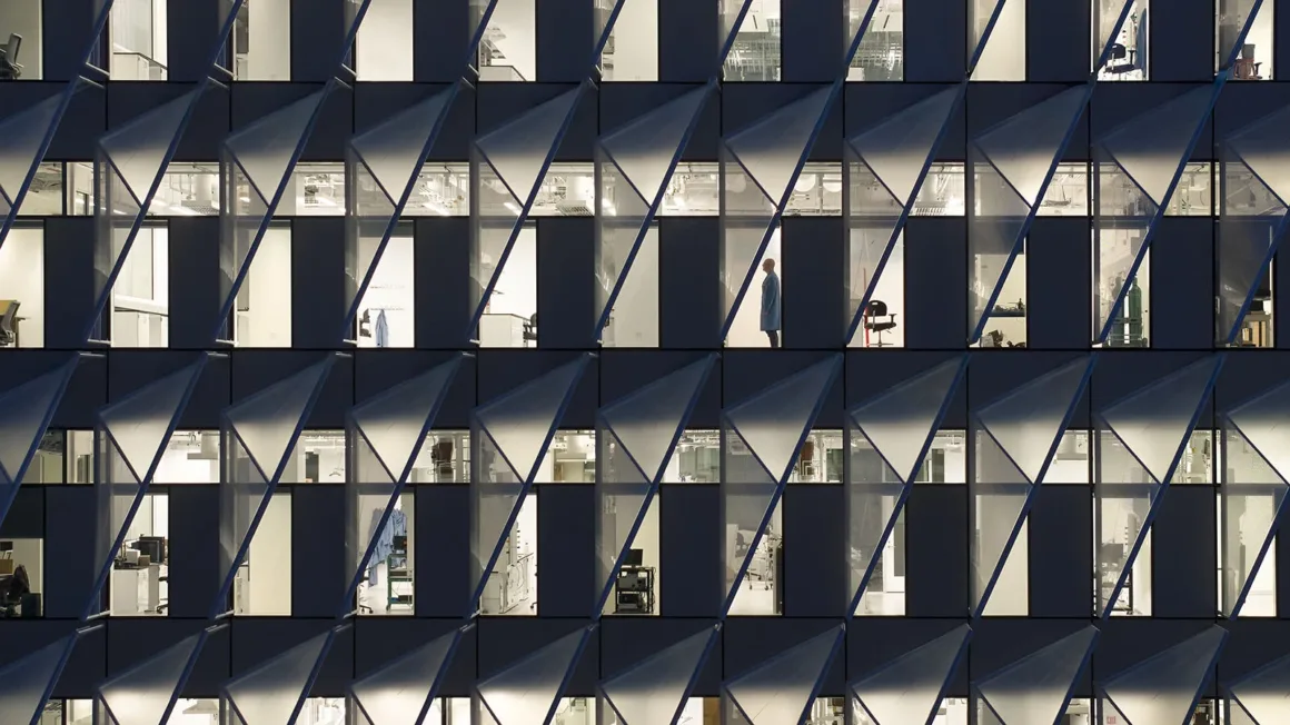 Architecture Photography MasterPrize 2026 Kazananları Açıklandı 29 Aydınlatılmış ofis veya laboratuvar pencerelerinden oluşan ızgara cephe, açılı yarı saydam üçgen paneller tarafından kısmen örtülüyor.