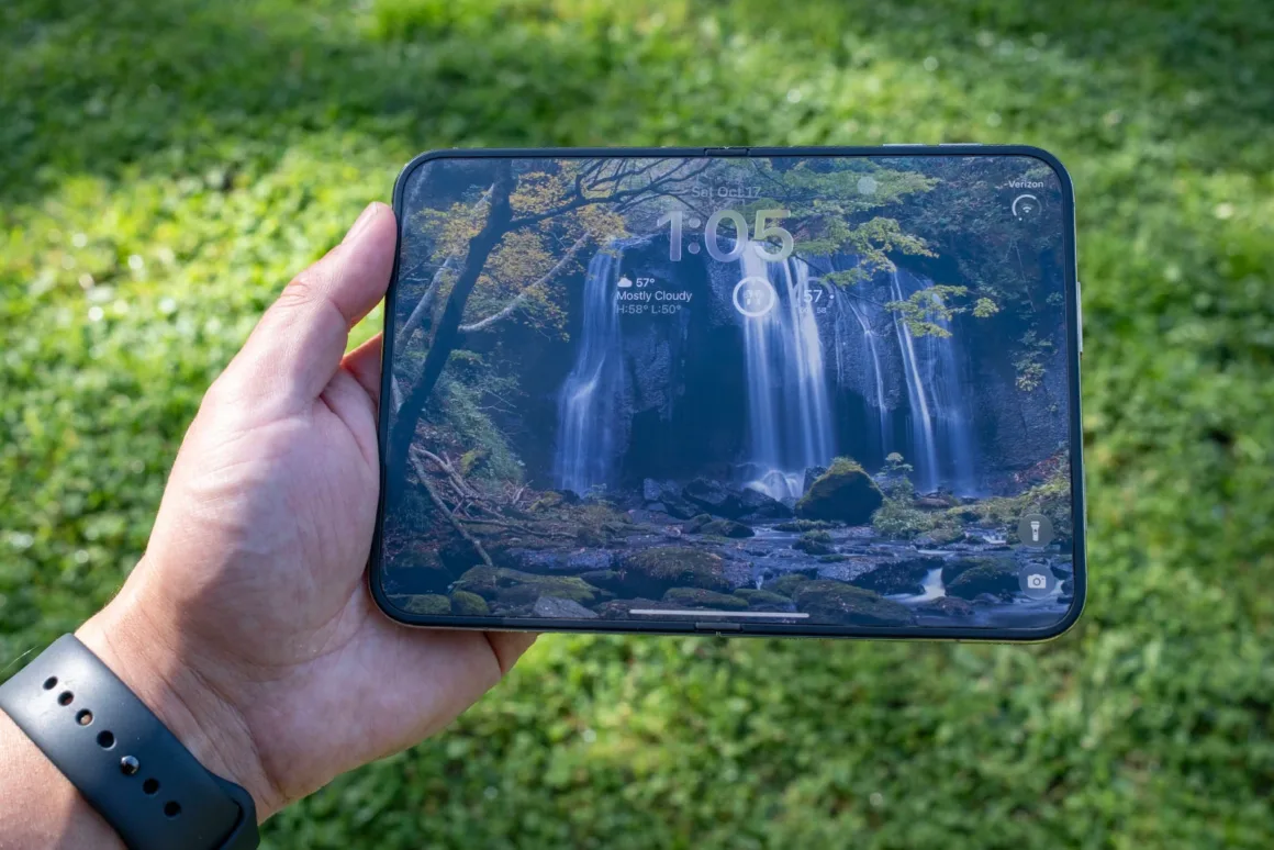 iPhone Duo İncelemesi: Etkileyici Tasarım, Kaçınılmaz Tavizler 19 Bir el, şelale duvar kâğıdı, saat 1:05 ve hava durumu araç takımlarını gösteren katlanabilir bir akıllı telefonu tutuyor.