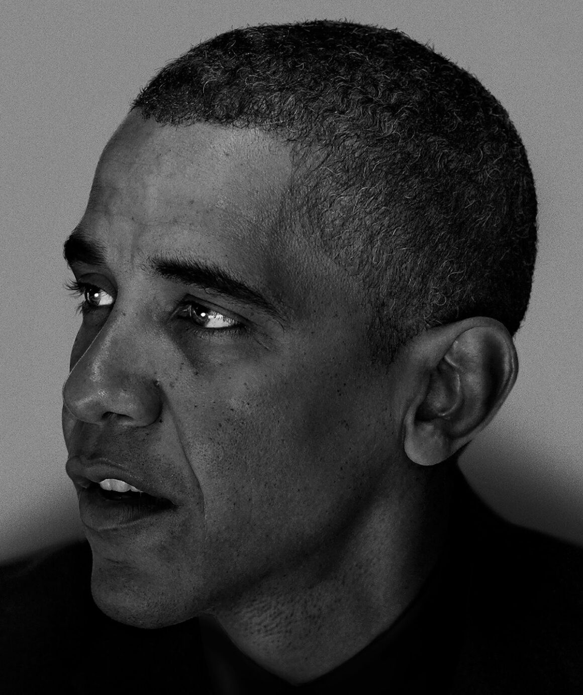 Barack Obama'nın yakın plan siyah-beyaz portresi; gözleri sola bakıyor ve ağzı, konuşmanın ortasındaymış gibi hafifçe açık. Arka planda sade gri bir fon bulunuyor.