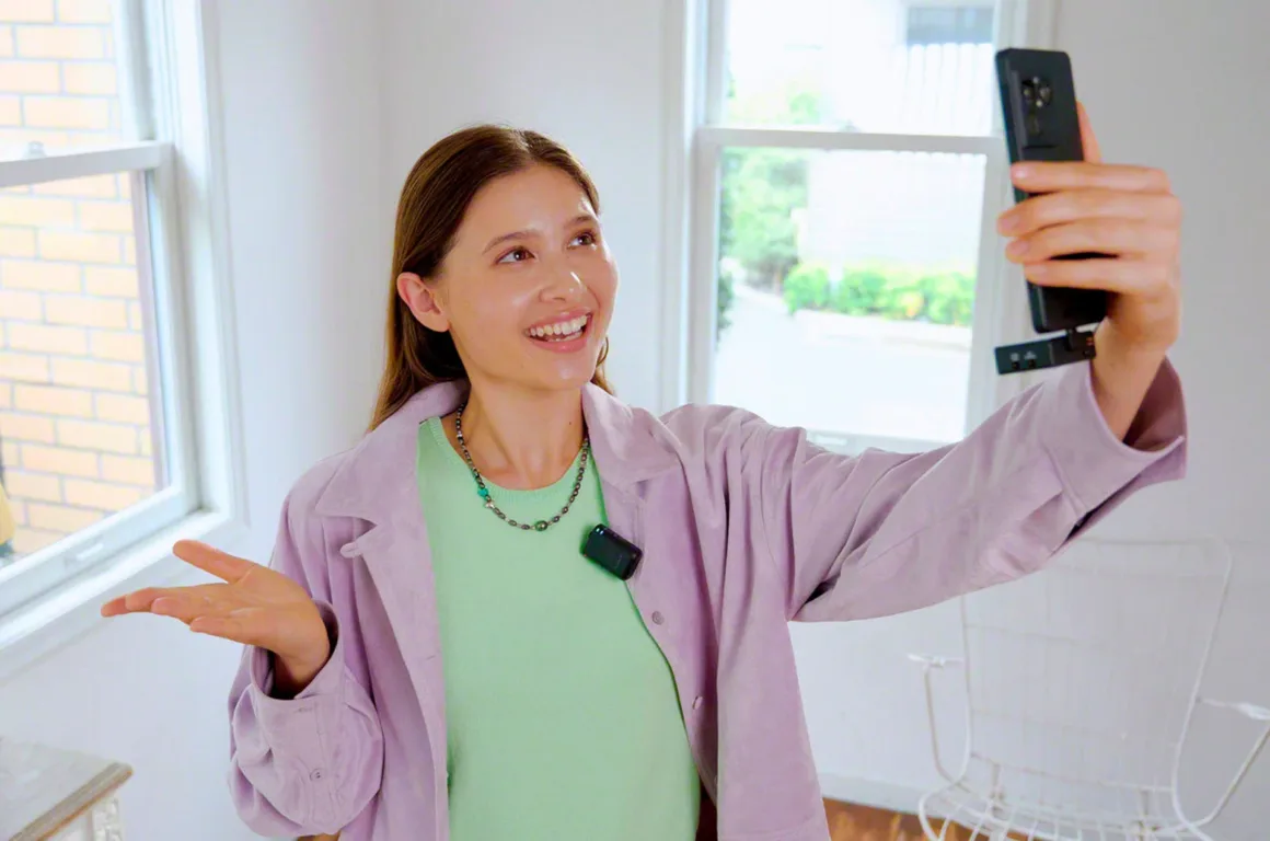 Sony ECM-W5: 32-Bit Float ve 500 Metre Menzilli Kablosuz Mikrofon Sistemi 26 A smiling woman in a light purple jacket and green shirt holds a smartphone up to take a selfie while wearing a small black microphone clipped to her collar.
