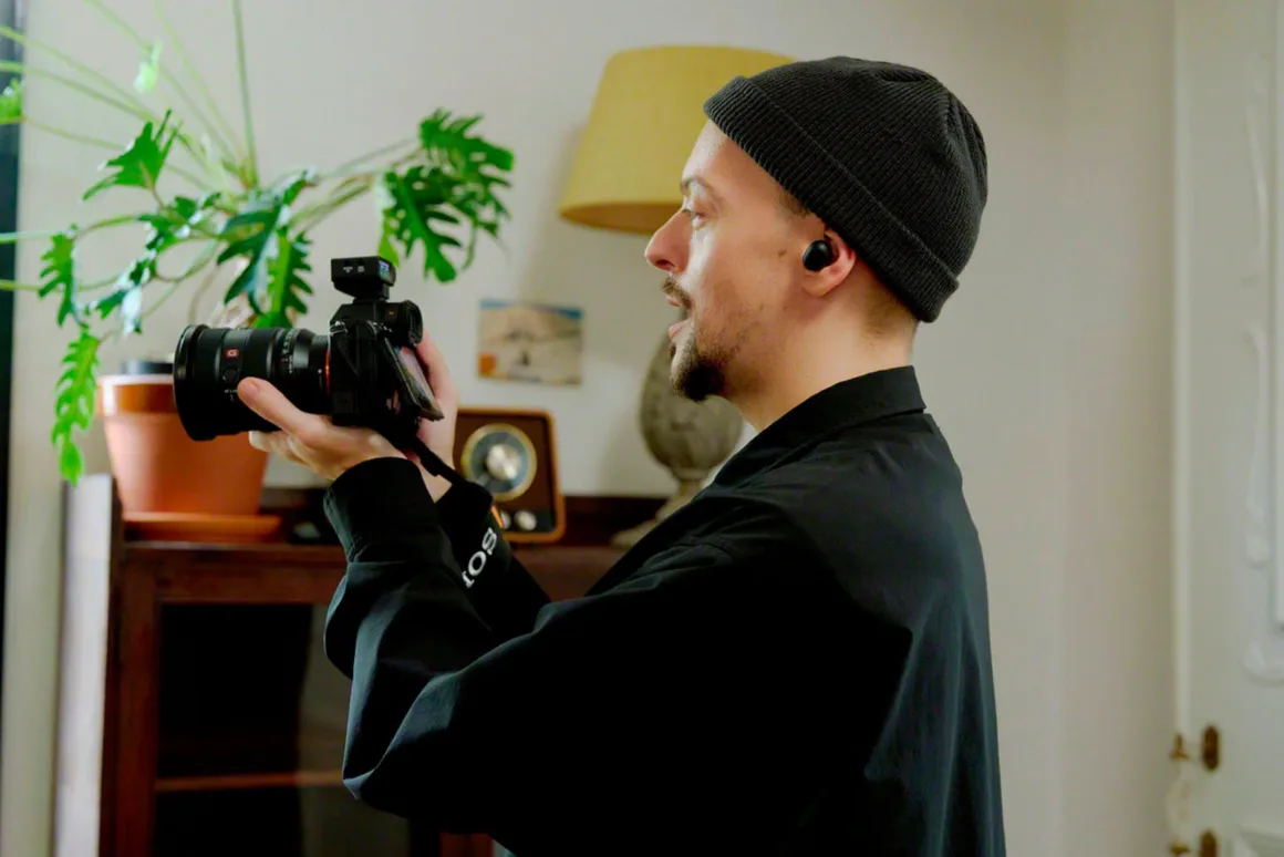 Sony ECM-W5: 32-Bit Float ve 500 Metre Menzilli Kablosuz Mikrofon Sistemi 27 A man wearing a black beanie and black shirt holds a camera up to his eye while standing in a room with a potted plant.