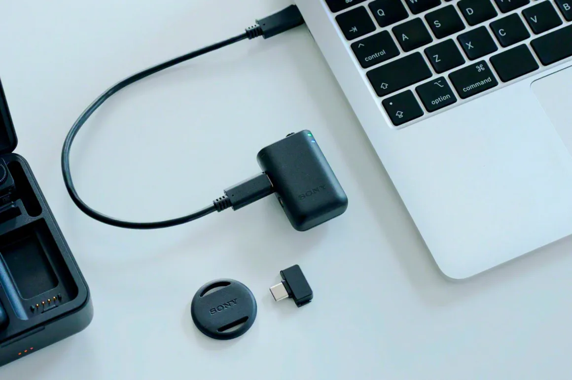 Sony ECM-W5: 32-Bit Float ve 500 Metre Menzilli Kablosuz Mikrofon Sistemi 23 Sony kablosuz mikrofon alıcısı USB-C kablosuyla bir dizüstü bilgisayara bağlı; masada yuvarlak manyetik bağlantı aparatı ve USB-C adaptörü bulunuyor.