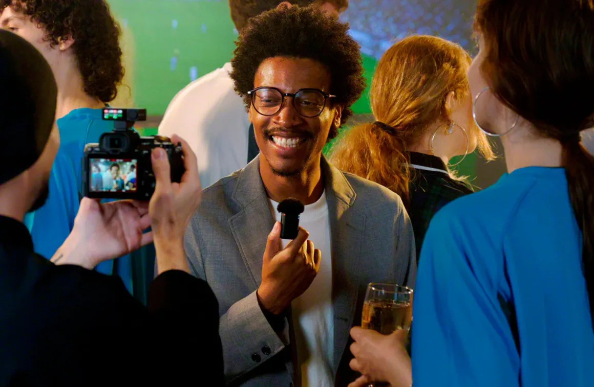 Sony ECM-W5: 32-Bit Float ve 500 Metre Menzilli Kablosuz Mikrofon Sistemi 24 A smiling man in a grey blazer holds a small microphone while being filmed by a person holding a camera in a crowded room.