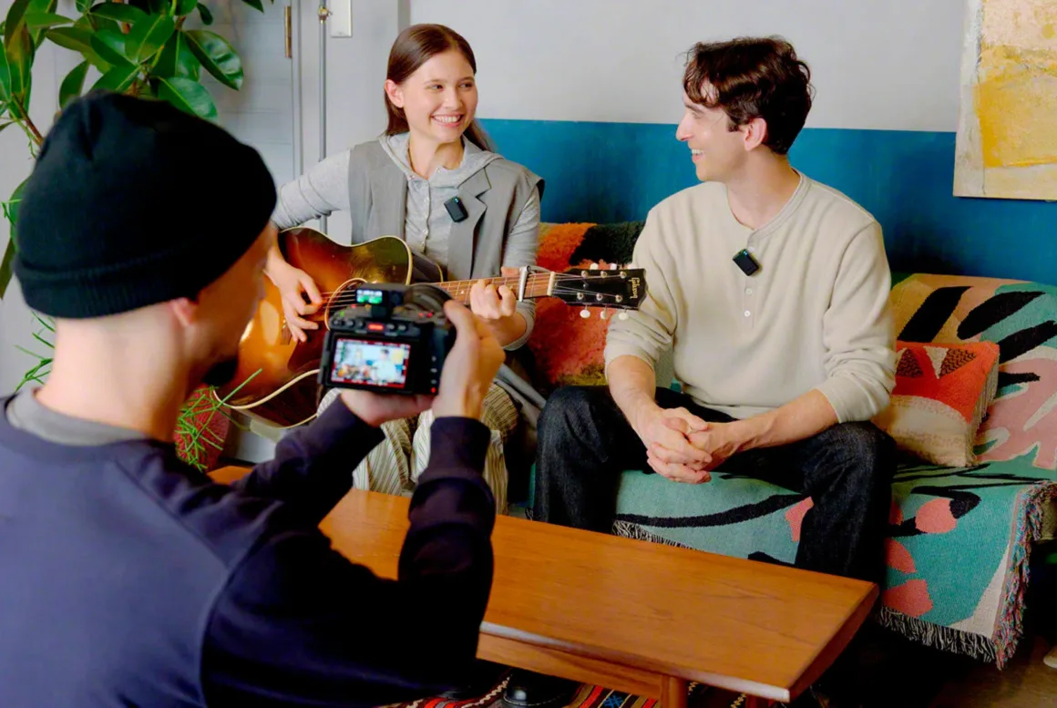 Sony ECM-W5: 32-Bit Float ve 500 Metre Menzilli Kablosuz Mikrofon Sistemi 25 A person in a black beanie films a woman playing an acoustic guitar and a man sitting on a couch, both wearing small microphones.