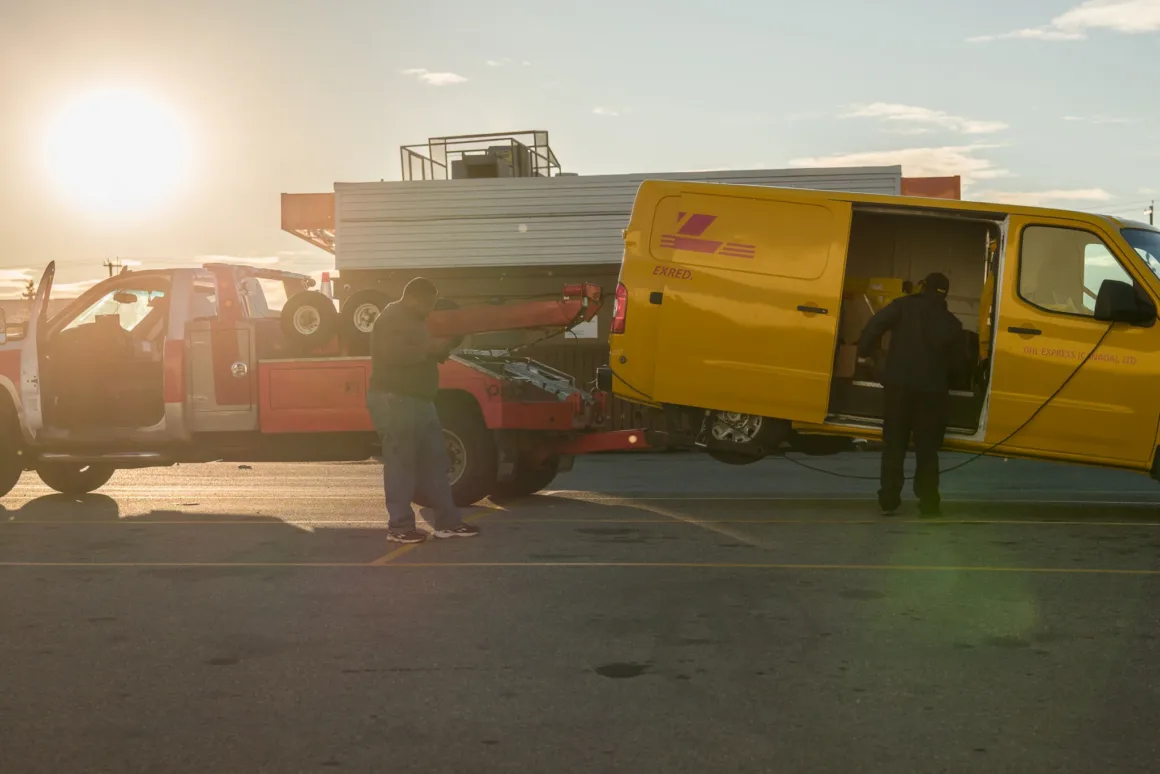 Sigma FP2 İncelemesi: Tuhaf Tasarım, Güçlü Hibrit Performans 34 Gün batımında bir otoparkta çekici araç sarı DHL Express minibüsüne bağlanmış, yakınlarda iki kişi duruyor.