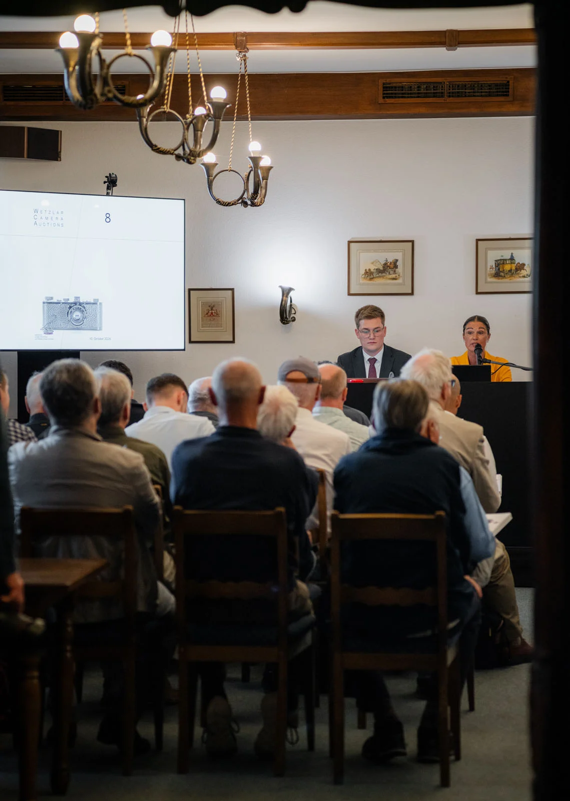 1923 Üretimi Leica 0-Series, 4,25 Milyon Euroya Satıldı 20 An audience sits in a room facing a stage where a man and a woman sit at a desk in front of a large screen displaying a camera graphic.