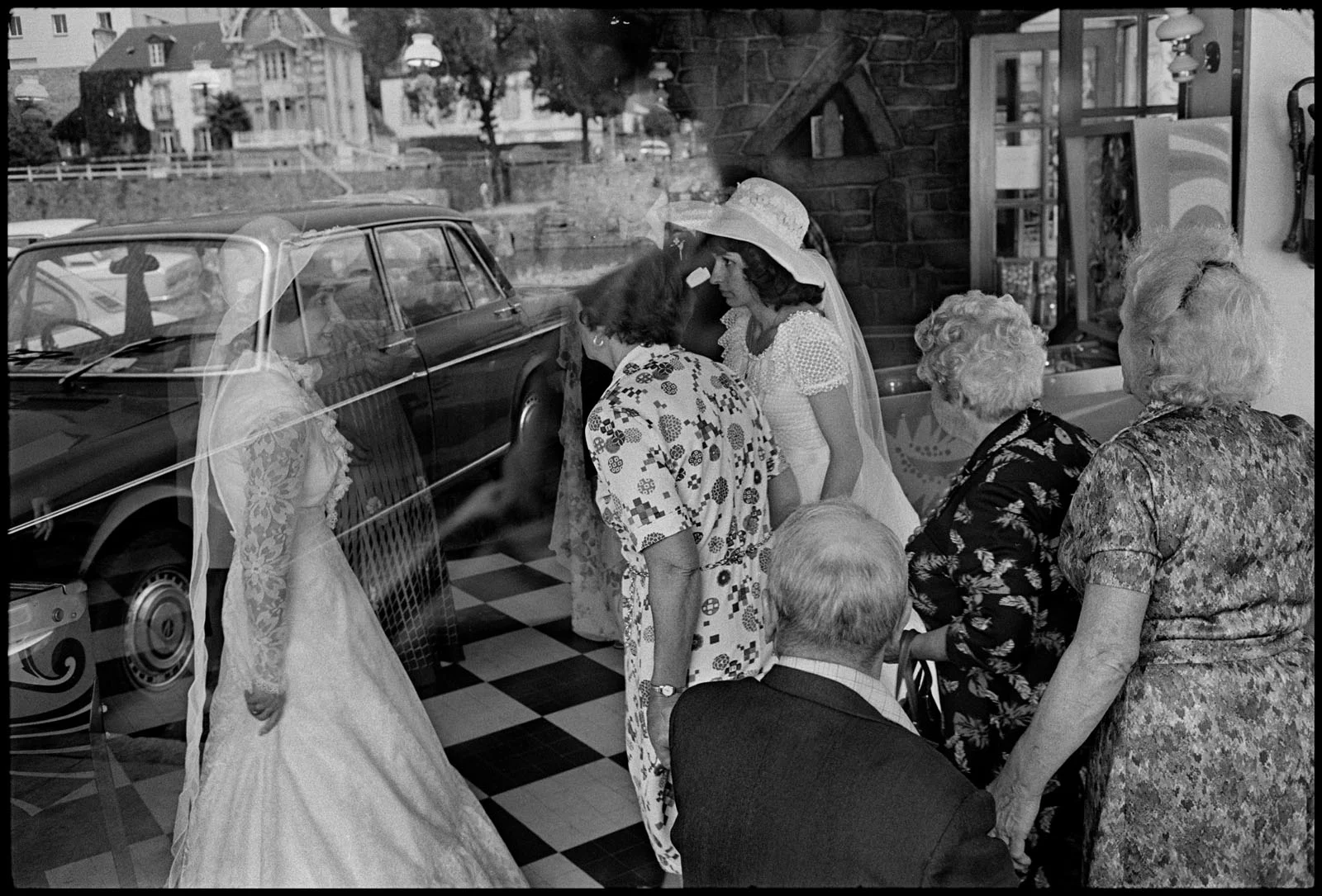 Magnum Fact/Fiction Square Print Sale 26 Ekim’de Başlıyor 27 A bride in a white gown and veil stands outside a window, reflected in the glass, while a group of people inside look out at her.