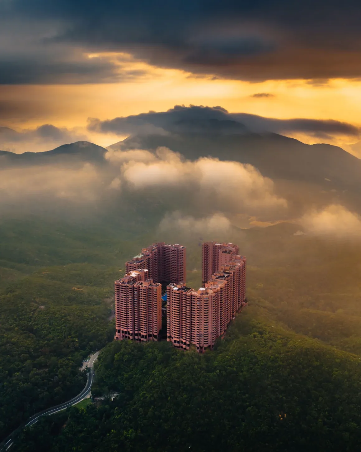 Architecture Photography MasterPrize 2026 Kazananları Açıklandı 25 U biçimindeki büyük pembe konut kompleksi, dramatik bulutlu gün batımı altında yemyeşil dağların arasında havadan görülüyor.