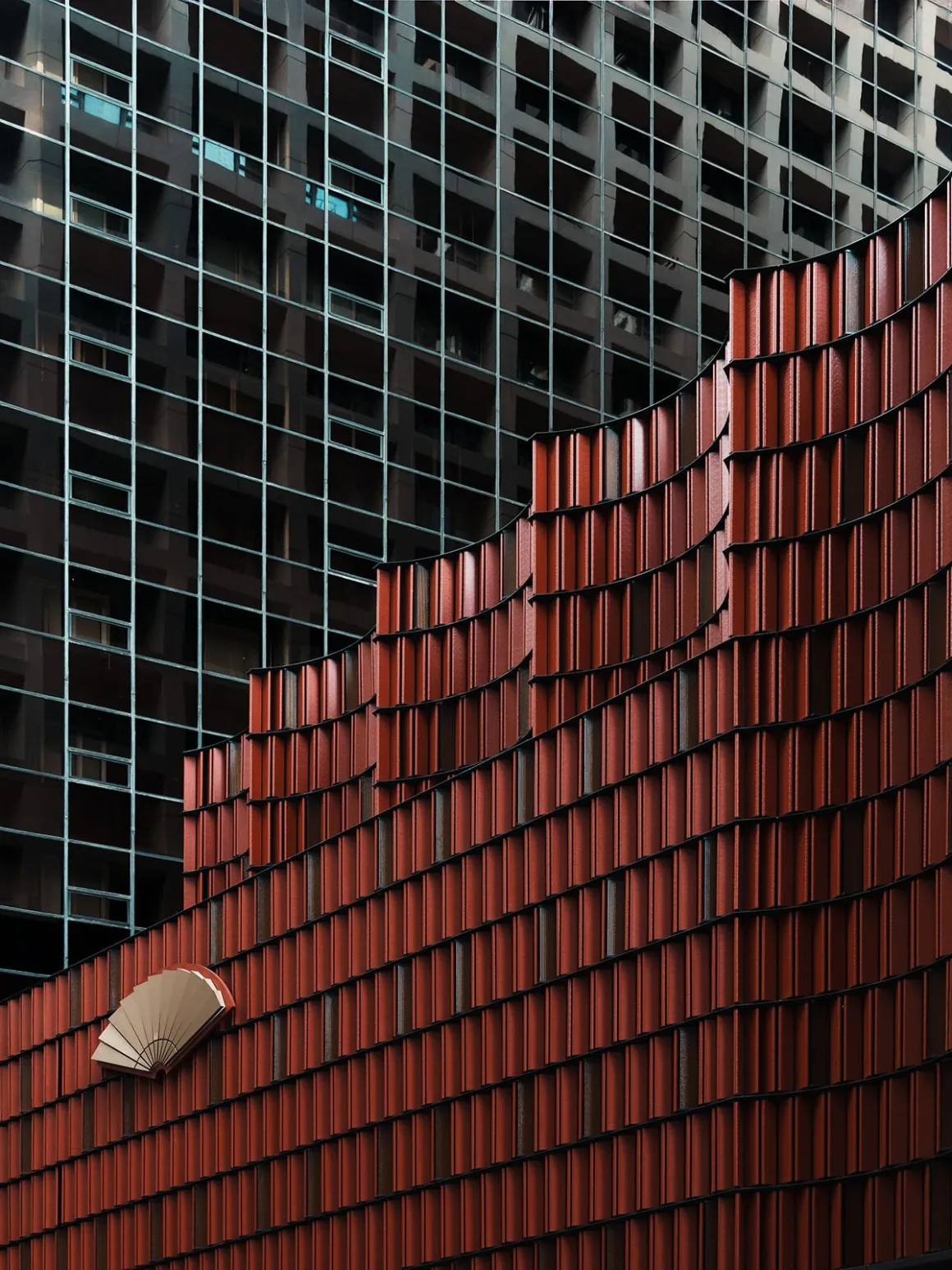 Architecture Photography MasterPrize 2026 Kazananları Açıklandı 24 Dikey elemanlarla biçimlendirilmiş terakota renkli kıvrımlı modern bina cephesi, koyu renkli ızgara desenli gökdelenin önünde görülüyor.