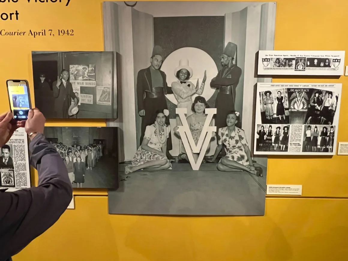 Charles “Teenie” Harris, Pittsburgh Walk of Fame’de Onurlandırıldı 23 Sarı duvar üzerindeki müze sergisinde Double V kampanyasına ilişkin tarihî siyah-beyaz fotoğraflar ve gazete kupürleri yer alıyor; bir ziyaretçi sergiyi telefonuyla fotoğraflıyor.