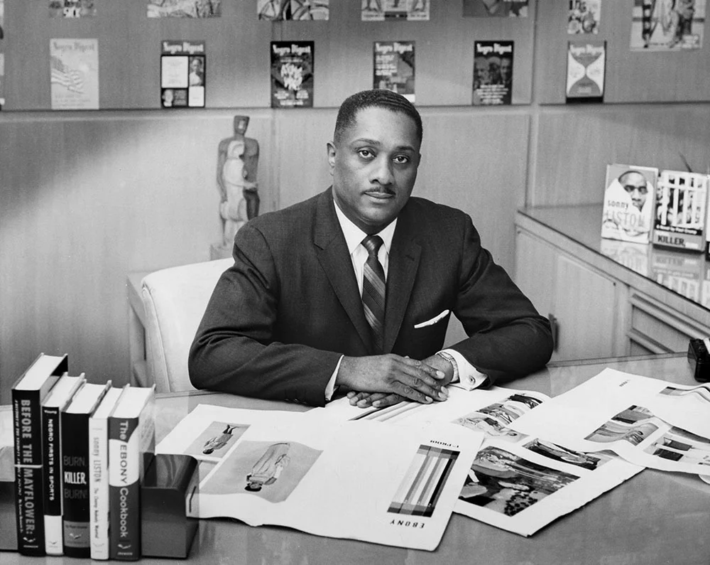 John H. Johnson, takım elbise ve kravatıyla çalışma masasının başında oturuyor; önünde kitaplar ve dergi prova baskıları, arka planda yayınlarla dolu raflar bulunuyor.