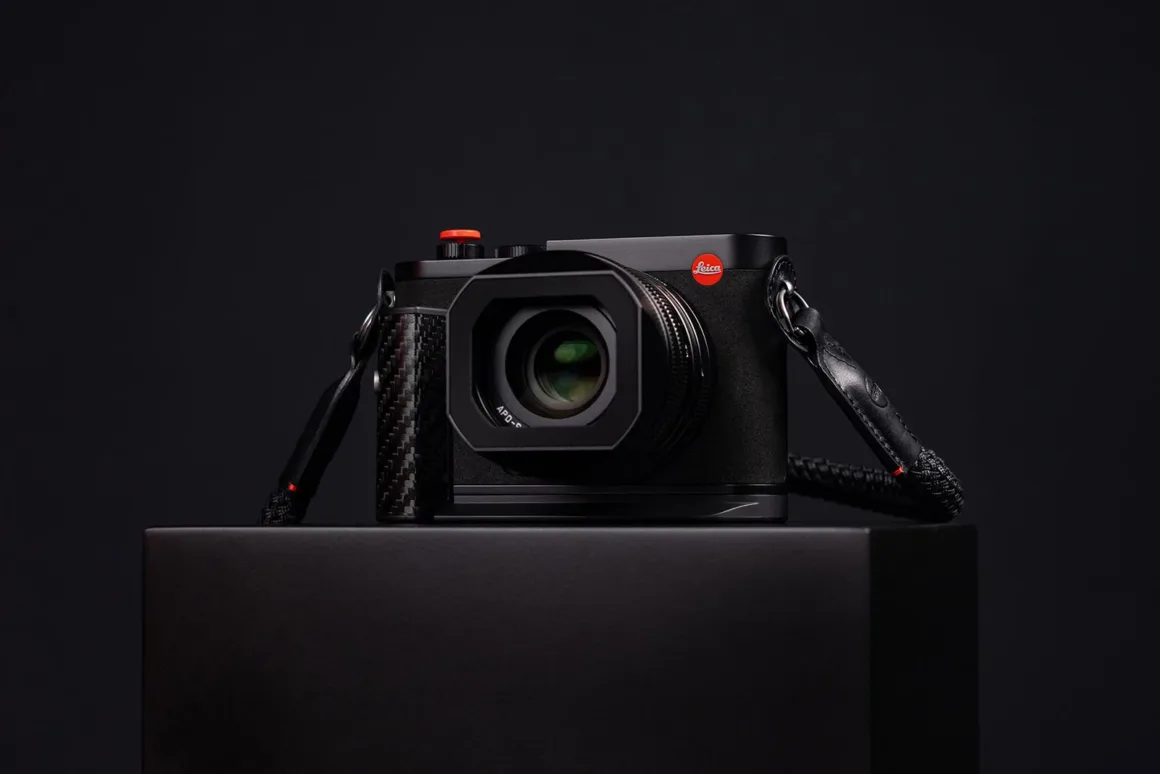 Leica Q3 43 Formula 1 Edition: Karbon Fiber Detaylı Özel Seri 19 Karbon fiber tutma bölümüne ve turuncu-kırmızı deklanşör düğmesine sahip siyah Leica fotoğraf makinesi, koyu fonda siyah bir kaide üzerinde duruyor.