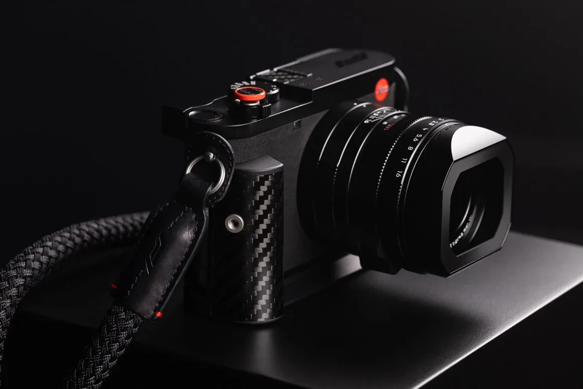 Leica Q3 43 Formula 1 Edition: Karbon Fiber Detaylı Özel Seri 21 Karbon fiber tutma bölümüne ve kırmızı deklanşör düğmesine sahip siyah dijital fotoğraf makinesi, örgülü siyah askısıyla koyu bir yüzey üzerinde duruyor.