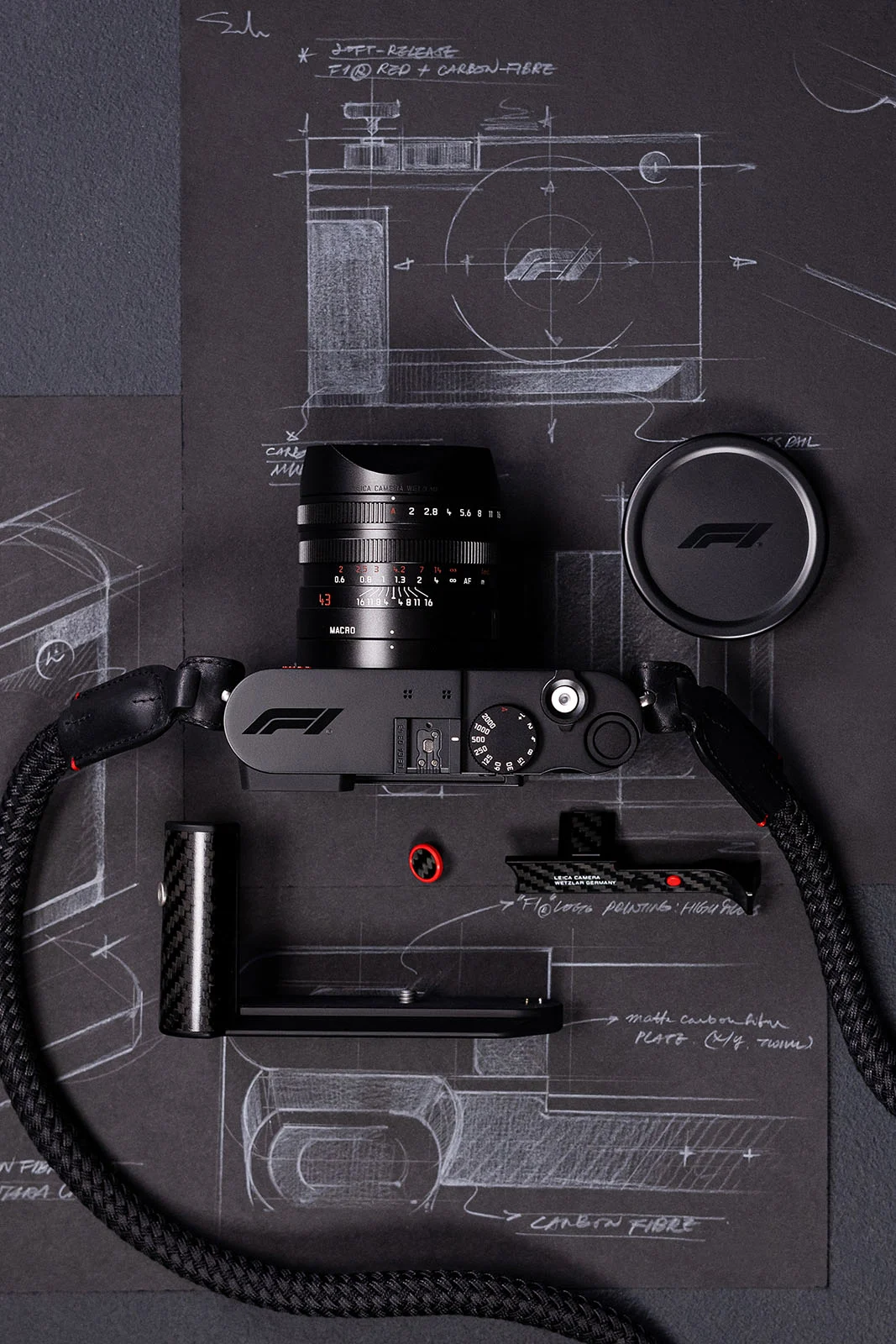 Leica Q3 43 Formula 1 Edition: Karbon Fiber Detaylı Özel Seri 22 F1 logolu siyah Leica fotoğraf makinesi, objektifi, objektif kapağı, tutma bölümü ve askısıyla birlikte teknik çizimler ve el yazısı notların bulunduğu koyu bir yüzey üzerinde üstten görüntüleniyor.