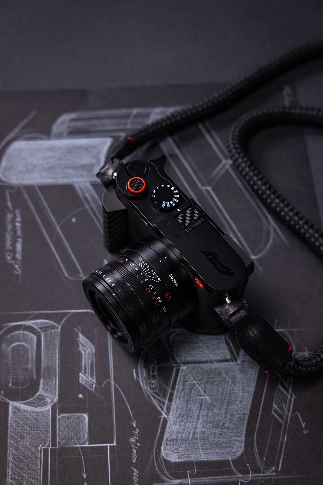Leica Q3 43 Formula 1 Edition: Karbon Fiber Detaylı Özel Seri 23 Kırmızı deklanşör düğmesine ve örgülü askıya sahip siyah dijital fotoğraf makinesi, beyaz araç teknik çiziminin bulunduğu koyu bir yüzey üzerinde duruyor.