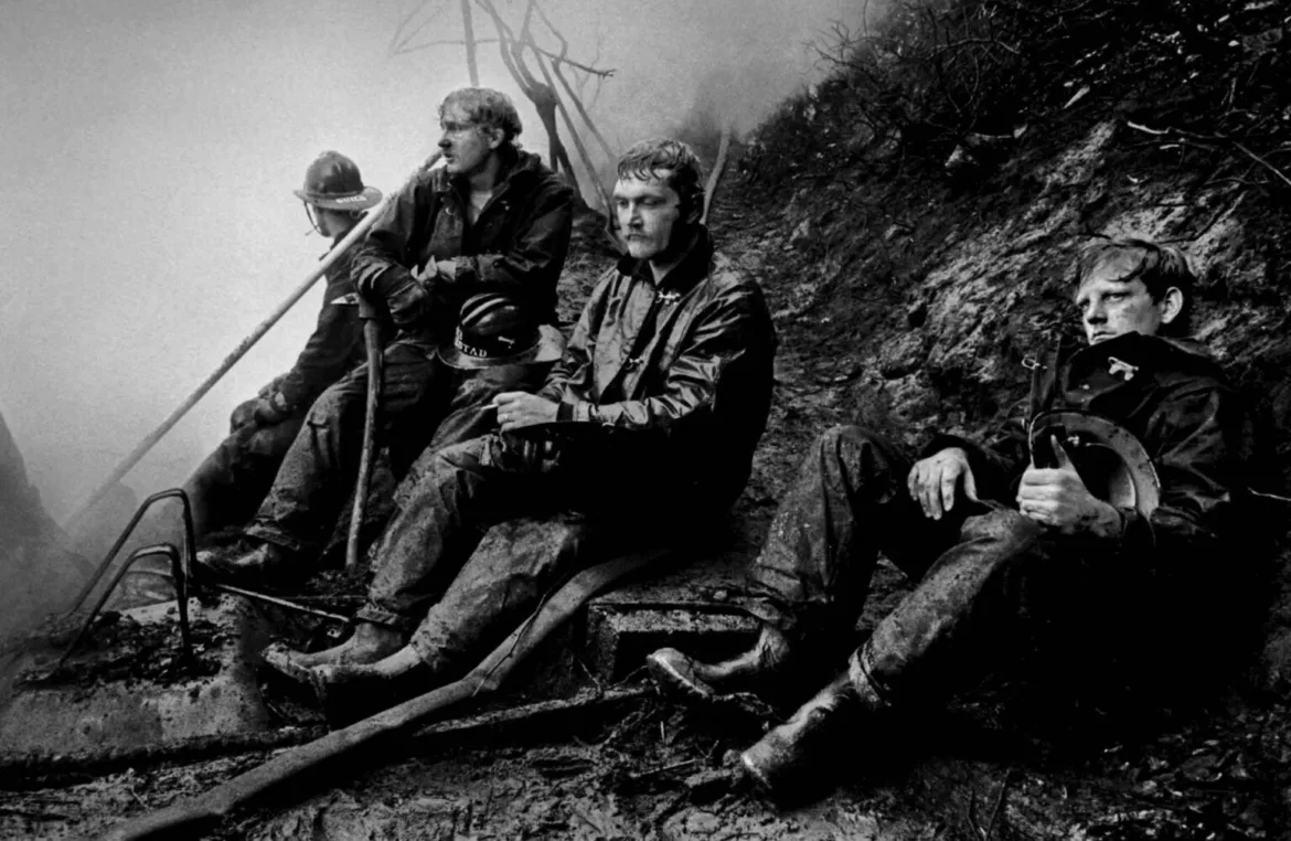 Four exhausted firefighters sit on a muddy, smoke-filled hillside, resting with their helmets and gear after battling a fire.