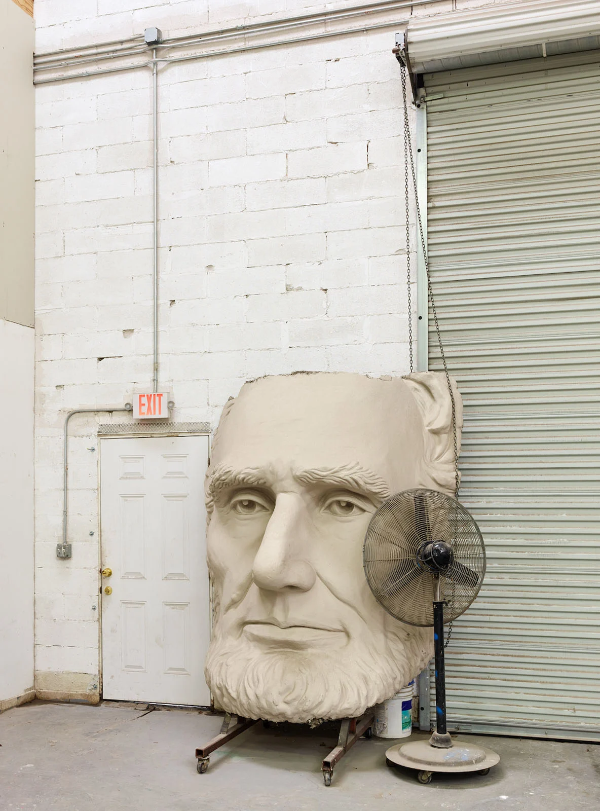 Magnum Fact/Fiction Square Print Sale 26 Ekim’de Başlıyor 23 A large, light-colored sculpture of Abraham Lincoln's head sits on a wheeled cart next to an industrial fan and a white exit door.