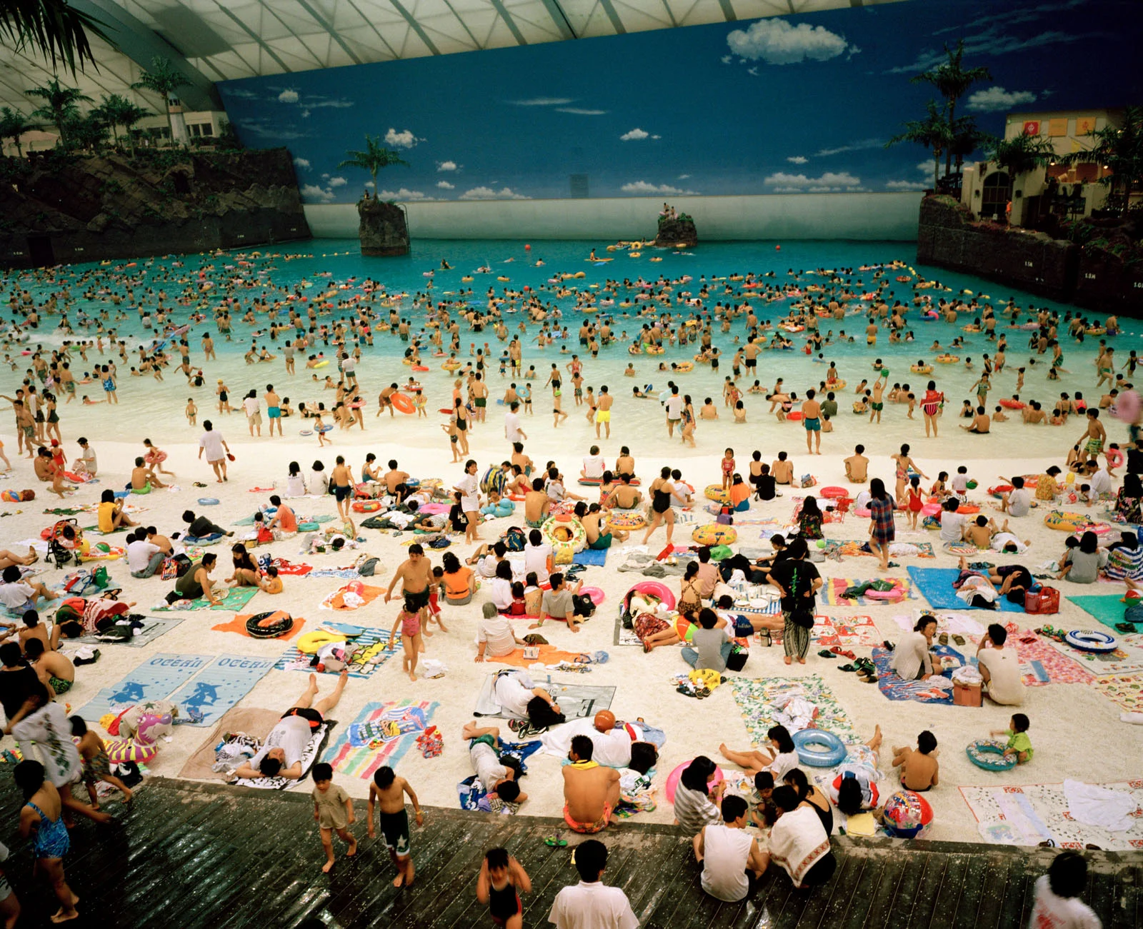 Magnum Fact/Fiction Square Print Sale 26 Ekim’de Başlıyor 19 A crowded indoor water park features a large wave pool with many swimmers and a sandy beach area filled with people sitting on towels and mats.