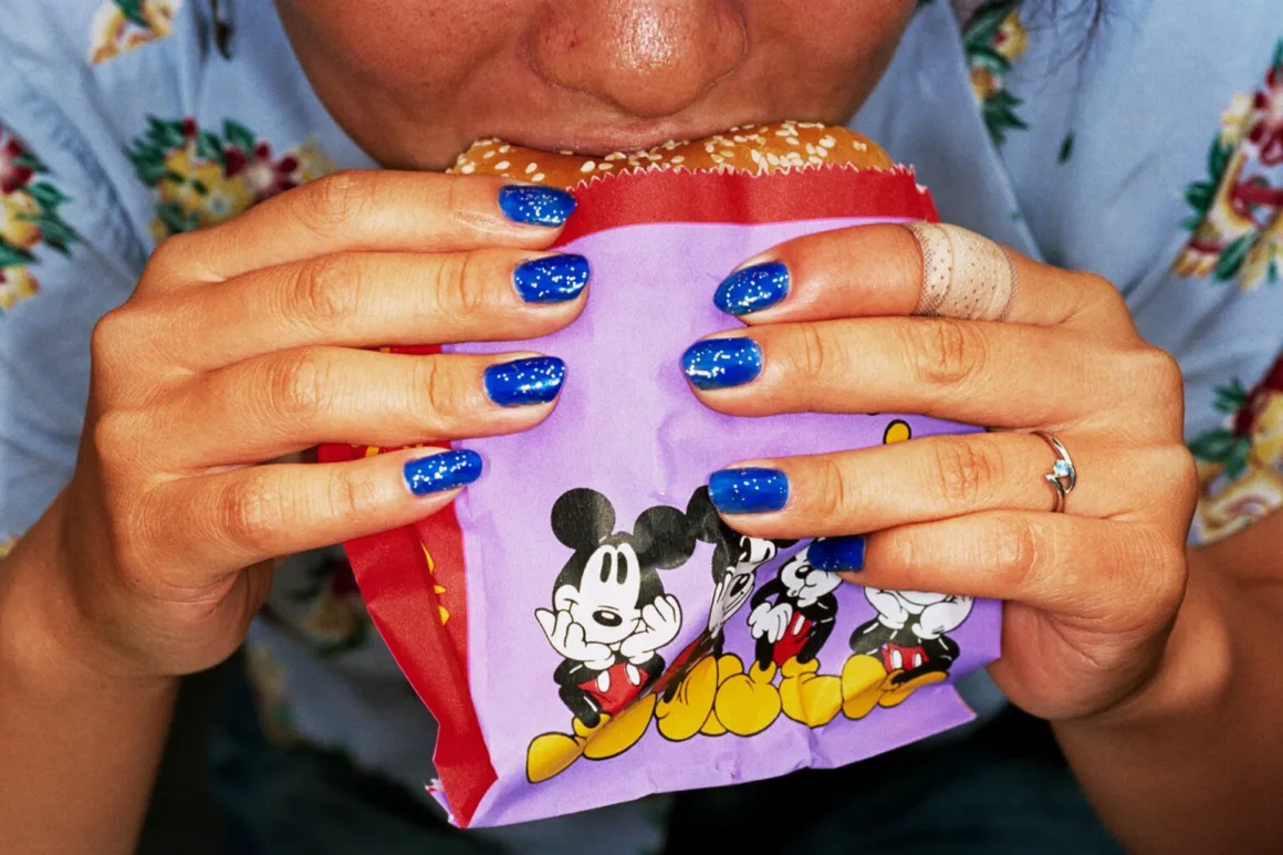Martin Parr’ın İkonik Common Sense Serisi 27 Yıl Sonra Yeniden Sergileniyor 1 Martin Parr’ın İkonik Common Sense Serisi 27 Yıl Sonra Yeniden Sergileniyor
Mavi simli ojeli bir kişi, mor Mickey Mouse ambalajına sarılı hamburgeri ısırırken tutuyor.