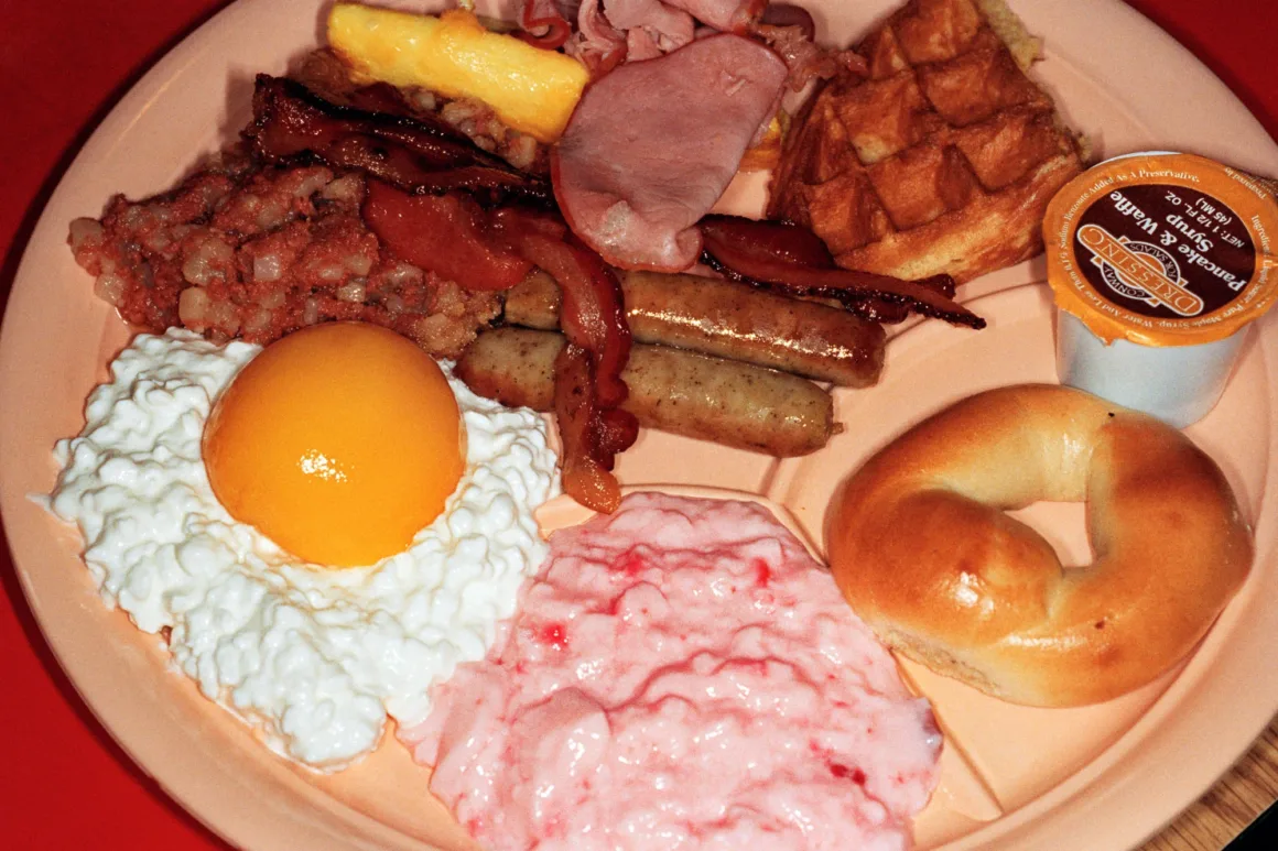 Martin Parr’ın İkonik Common Sense Serisi 27 Yıl Sonra Yeniden Sergileniyor 20 Bölmeli bir tabakta waffle, sosis, bacon, jambon, corned beef hash, bagel, şeftalili lor peyniri ve pembe yoğurttan oluşan bir kahvaltı bulunuyor.