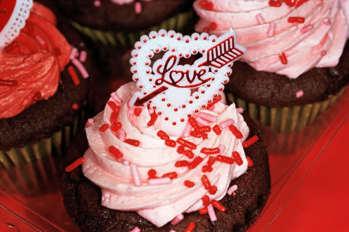 Martin Parr’ın İkonik Common Sense Serisi 27 Yıl Sonra Yeniden Sergileniyor 26 Pembe kremayla kaplanmış ve kırmızı şekerlemelerle süslenmiş çikolatalı cupcake'lerin üzerinde, okla delinmiş beyaz kalp biçimli 'Love' süsleri bulunuyor.