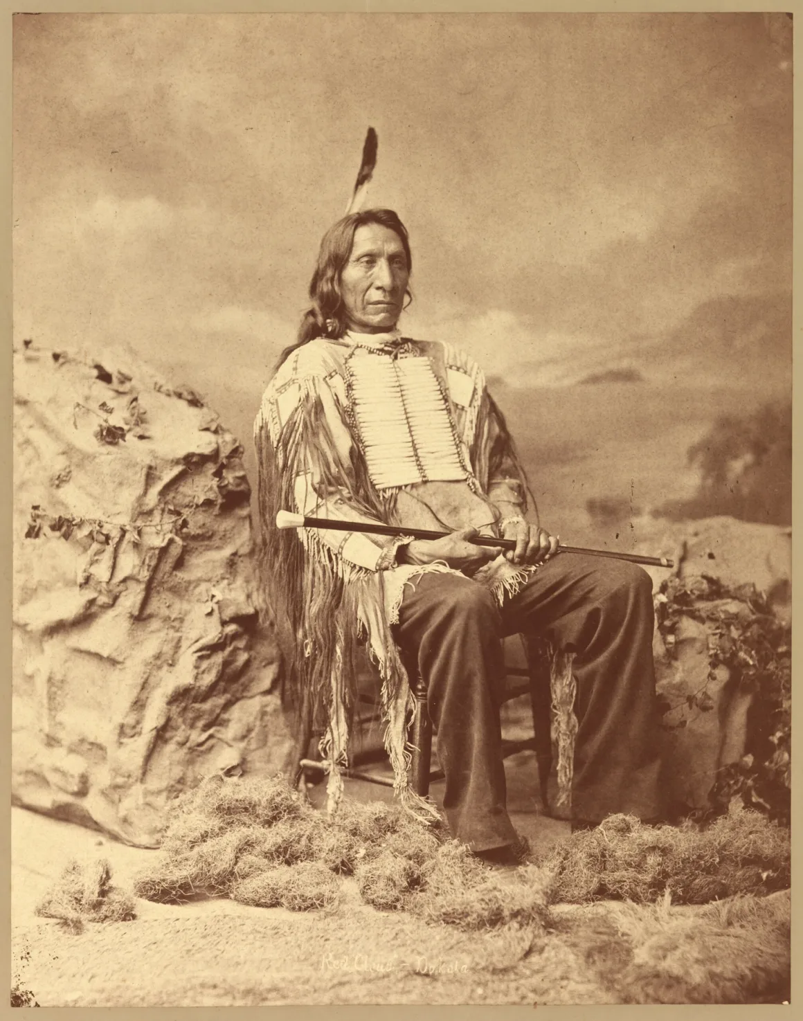 Lakota lideri Red Cloud, ahşap sandalyede oturup baston tutarken boyalı fon ve yapay bitkilerin önünde poz veriyor.
