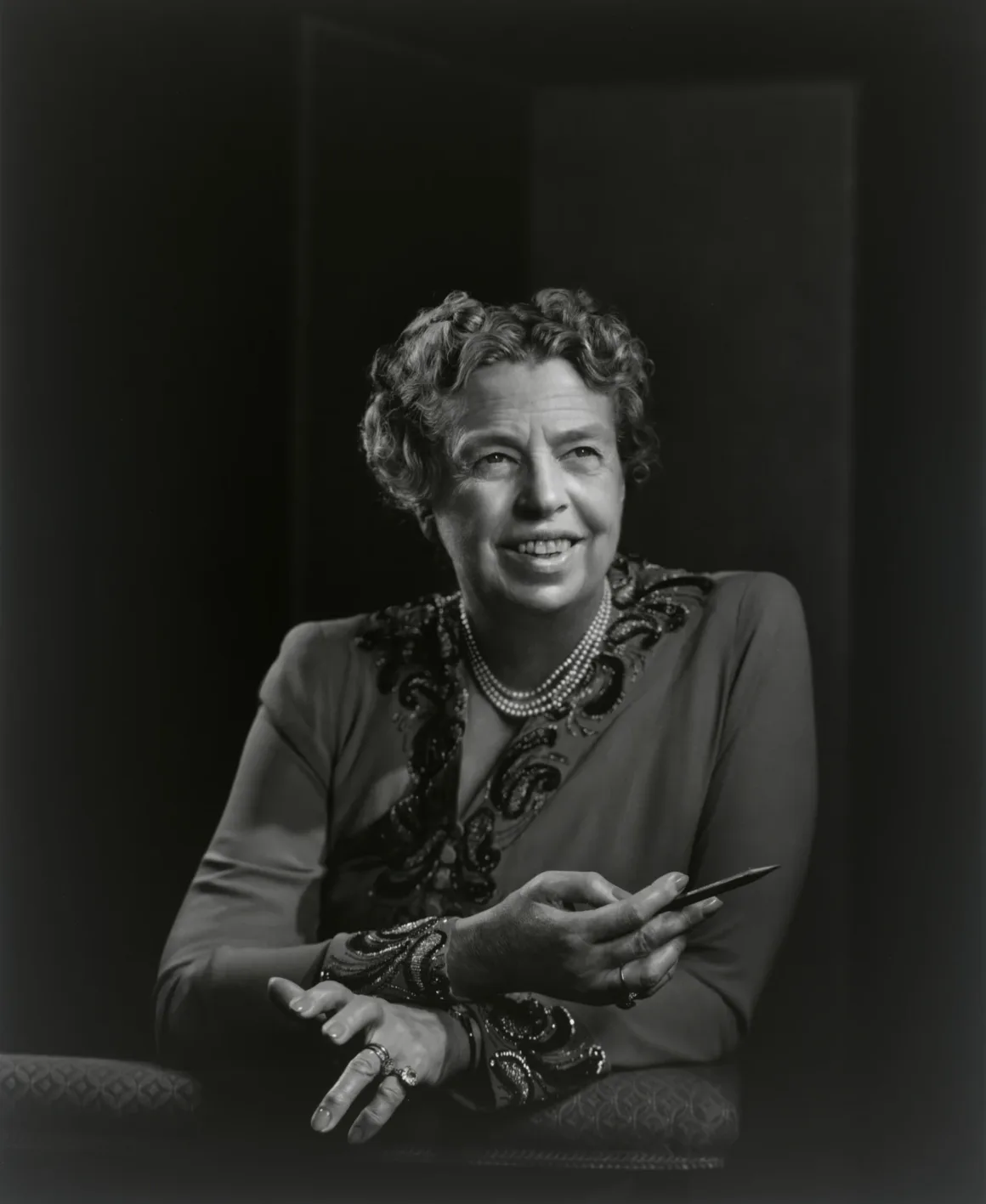 Eleanor Roosevelt, boncuk işlemeli elbise ve inci kolyeyle, elinde kalem tutarken yana doğru bakıyor.