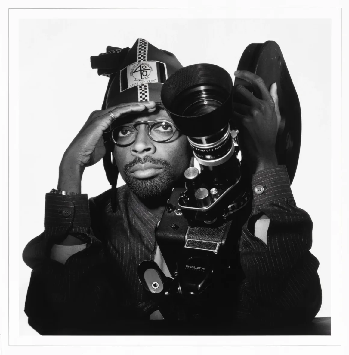 Spike Lee, markalı bir şapka ve gözlük takarak siyah-beyaz portrede poz veriyor ve elinde Bolex film kamerası tutuyor.