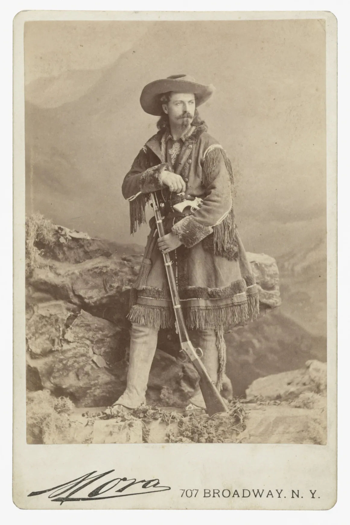 William “Buffalo Bill” Cody, püsküllü güderi ceketiyle kayalık bir dekor üzerinde ayakta duruyor ve elinde tüfek tutuyor.