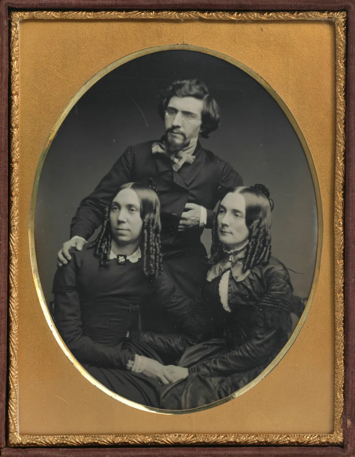 19. yüzyıl ortasına ait resmi kıyafetler içindeki iki oturan kadının arkasında duran sakallı bir adamı gösteren daguerreotype portre.