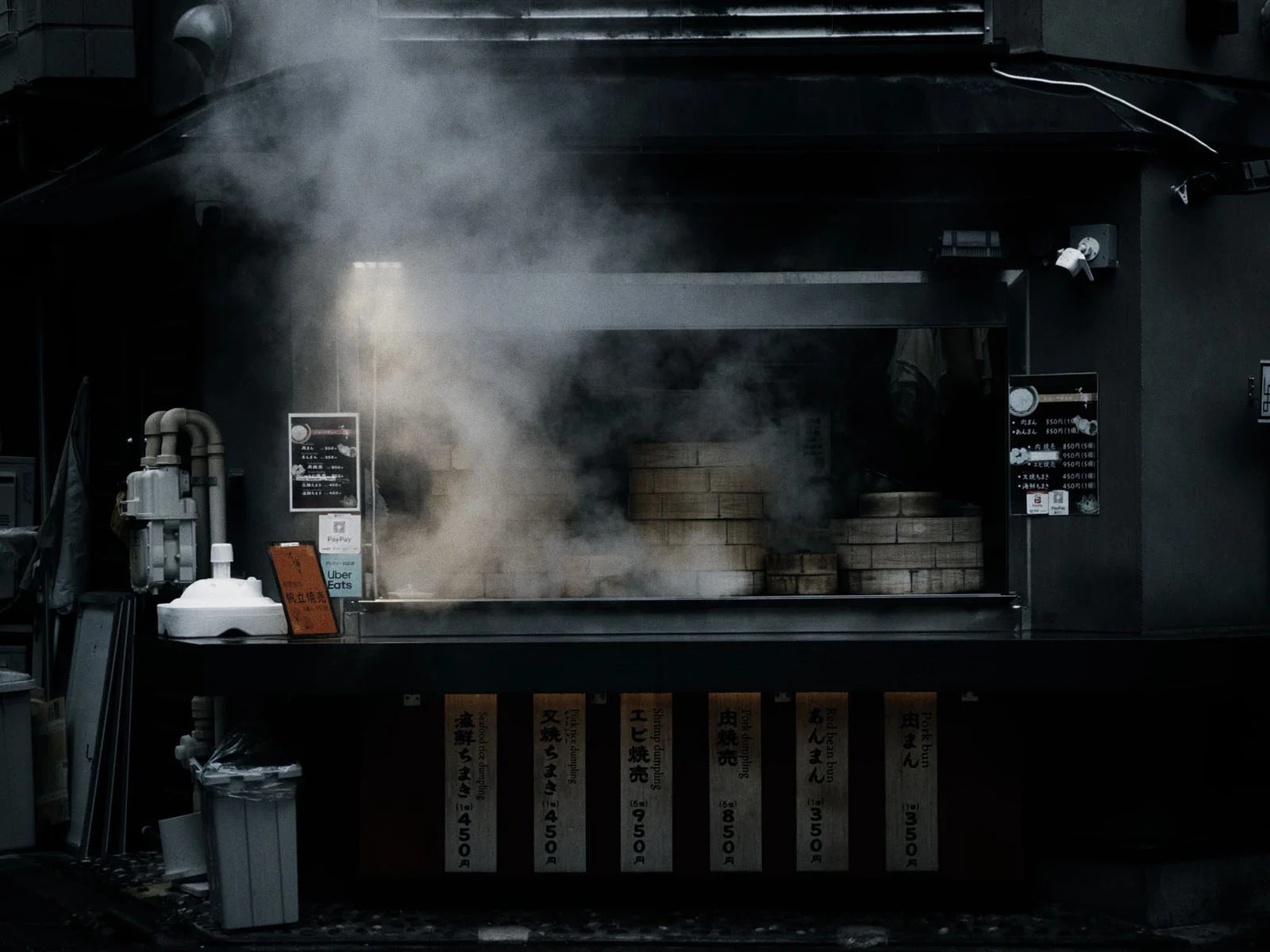 Halide 3.2, Apple’ın Yeni Değişken Diyaframını Daha da İleri Taşıyor 21 A street food stall with stacks of bamboo steamers emitting thick steam, featuring menu signs with Japanese text and English translations for various dumplings and buns.