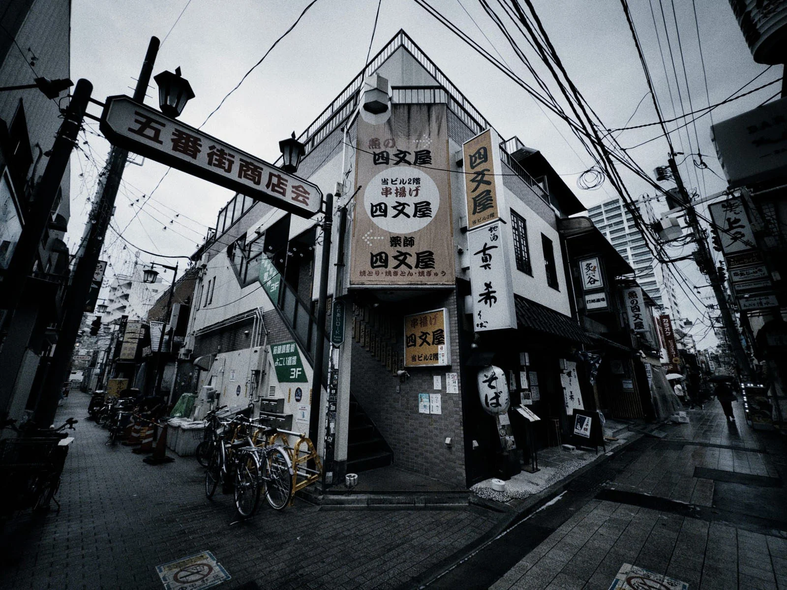 Halide 3.2, Apple’ın Yeni Değişken Diyaframını Daha da İleri Taşıyor 22 A street corner in Japan featuring a multi-story building with Japanese signage, a bicycle rack, and overhead power lines under a cloudy sky.
