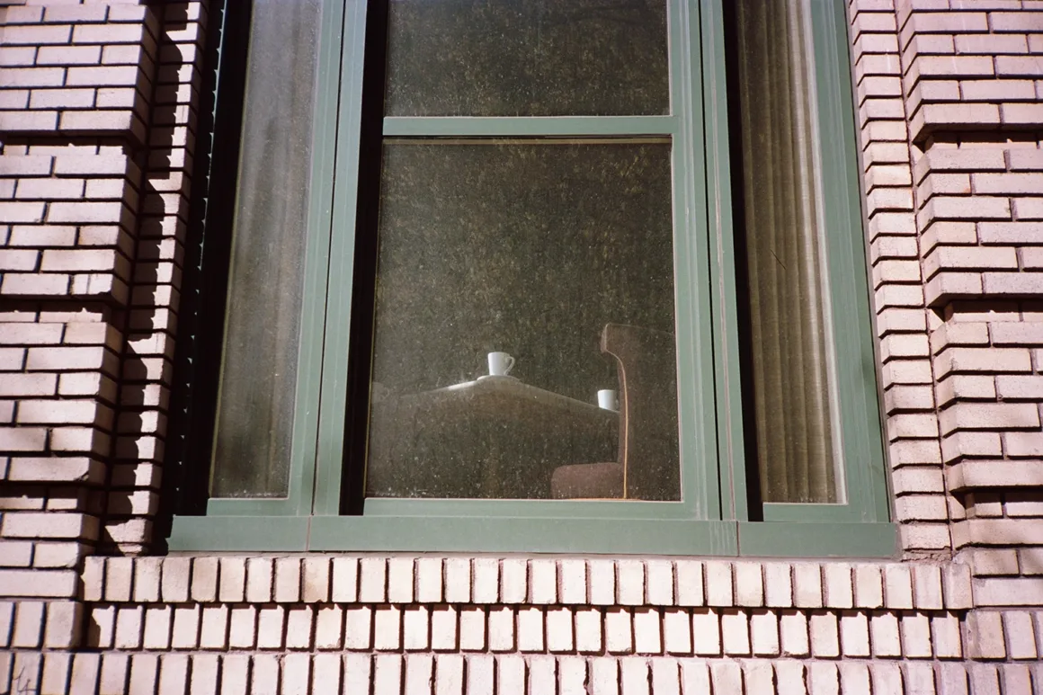 Kodak Portra 160: İnce Grenli Renkli Negatif Filmin Gücü 23 Tozlu bir pencerenin ardından tuğla bir yapının içinde iki beyaz kupanın bulunduğu masa ve bir sandalye görülüyor.