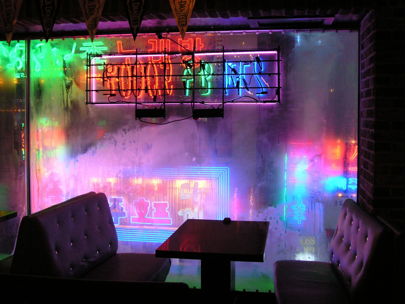 Magnum Fact/Fiction Square Print Sale 26 Ekim’de Başlıyor 21 A dimly lit booth with two dark, tufted chairs and a small table sits in front of a window covered in colorful neon signs.