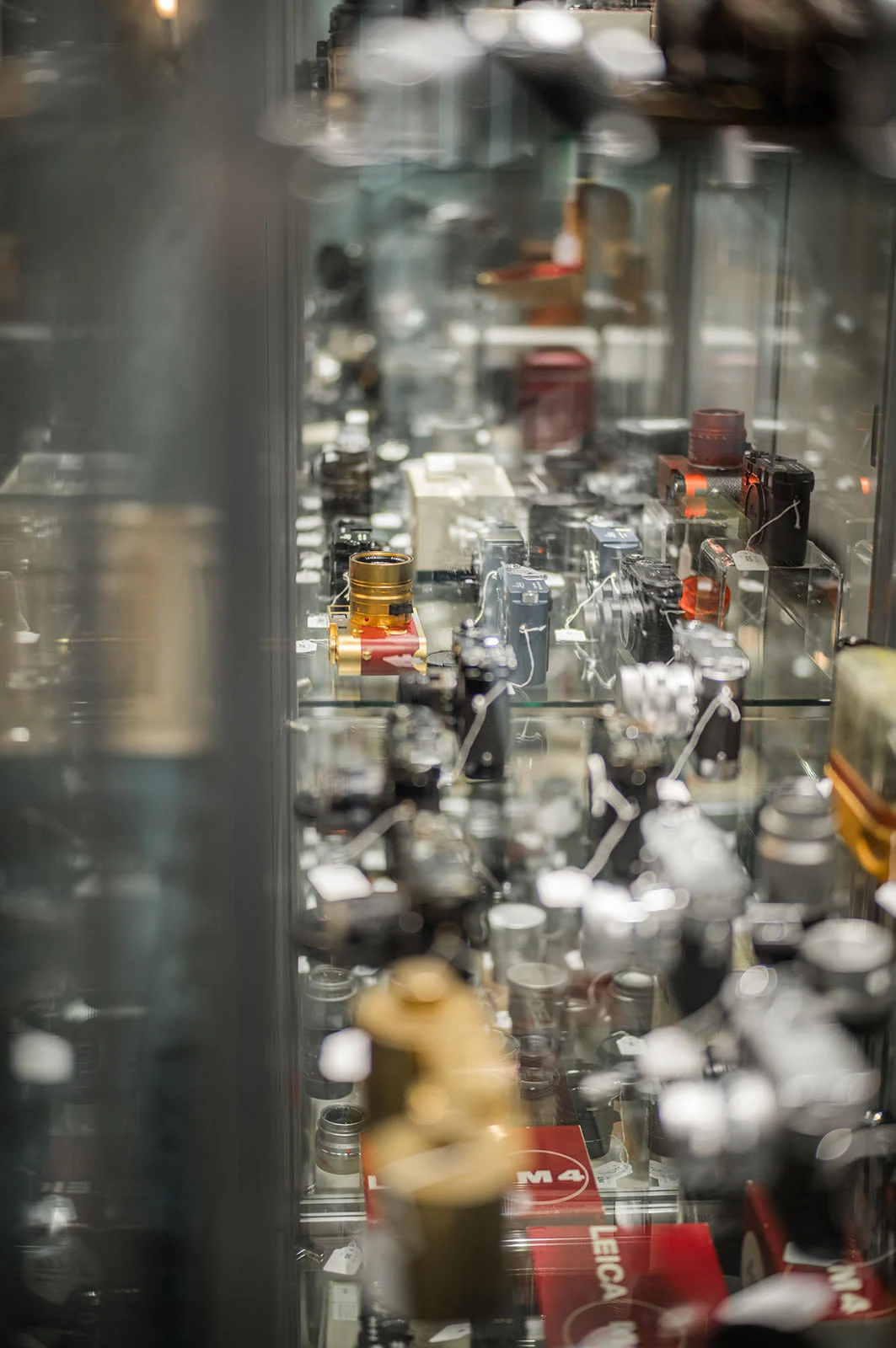 1923 Üretimi Leica 0-Series, 4,25 Milyon Euroya Satıldı 30 A display case filled with numerous vintage cameras and lenses on glass shelves, with red Leica M4 boxes visible at the bottom.